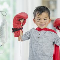 A young Asian boy is in indoors in a hospital. He is wearing casual clothes, facepaint, boxing gloves, and a cape. He is pretending to be a superhero fighting his disease. He looks very cheerful.